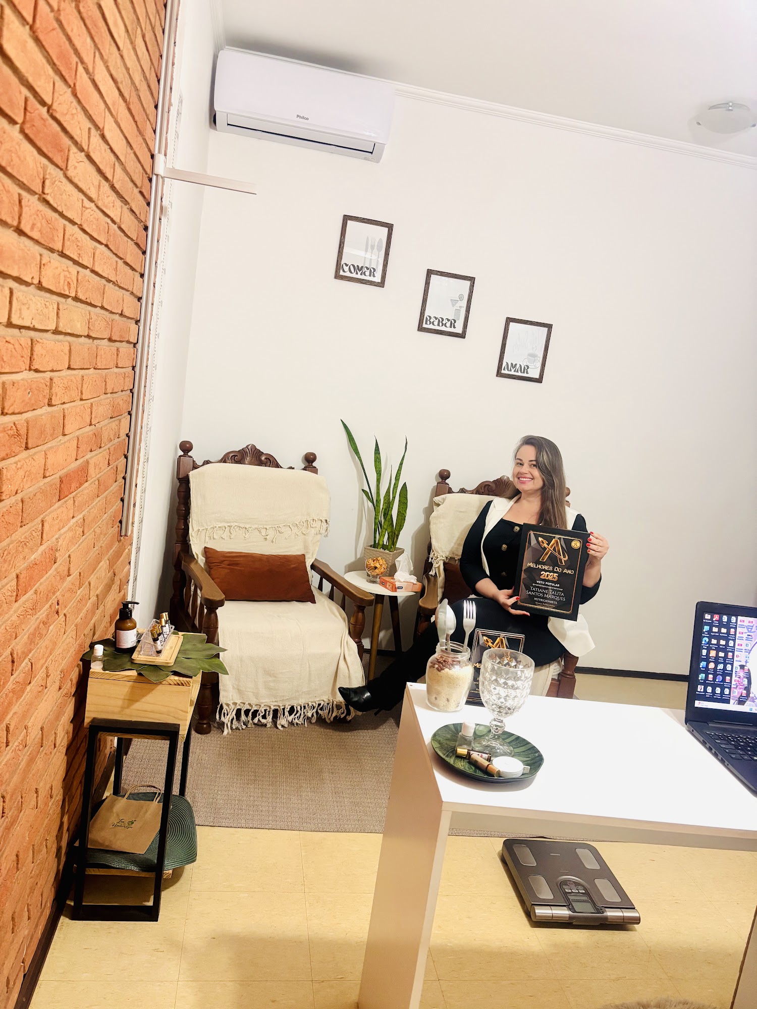 Tatiane Santos sentada em uma poltrona de madeira do consultório, ao lado da planta, sorrindo, com a outra poltrona e os quadros ao fundo